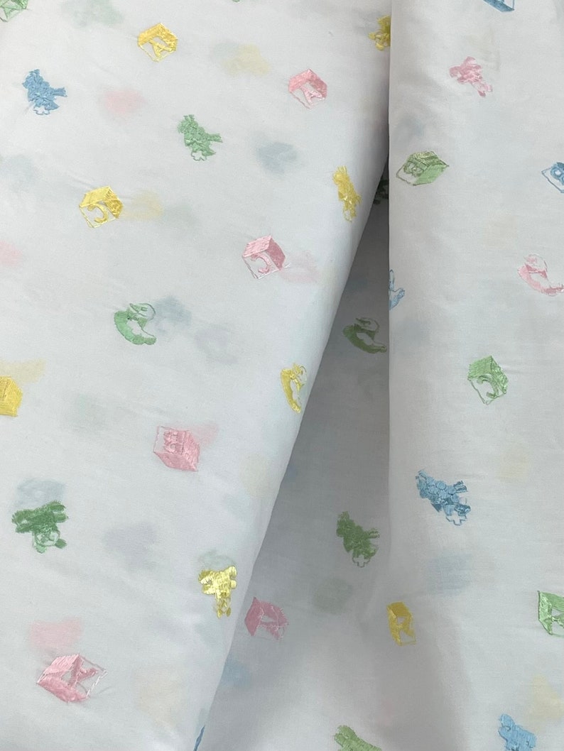 May include: White fabric with a repeating pattern of colourful embroidered toy blocks. The blocks are in shades of pink, yellow, green, and blue.