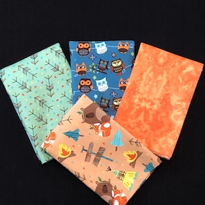 Puede incluir: Cuatro cuadrados de tela con diferentes patrones. Un cuadrado tiene un patrón verde y blanco de flechas y triángulos. Otro cuadrado tiene un patrón azul y blanco de búhos. Un tercer cuadrado tiene un patrón marrón y naranja de osos y zorros. El último cuadrado es de color naranja sólido.