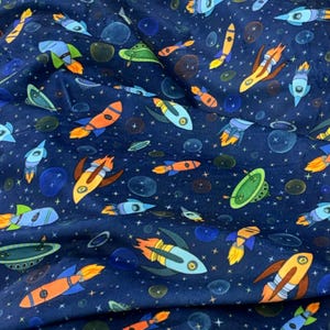 May include: Dark blue fabric with a repeating pattern of colorful rockets, planets, and stars. The rockets are in shades of orange, yellow, blue, and green. The planets are various sizes and colors, with white stars scattered throughout.