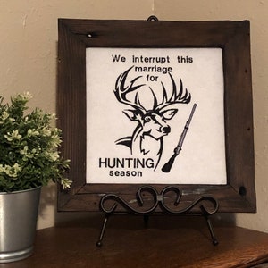 May include: A framed canvas with a black and white illustration of a deer and a rifle. The text on the canvas reads "We interrupt this marriage for hunting season".