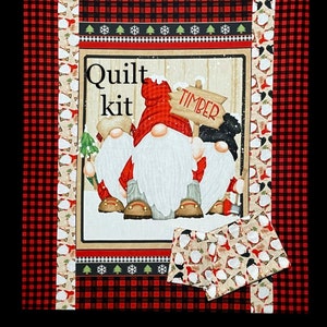 May include: Quilt kit featuring three gnomes with red hats and white beards. The gnomes are standing in front of a wooden sign that says "TIMBER". The background is a red and black checked fabric with a white snowflake border.