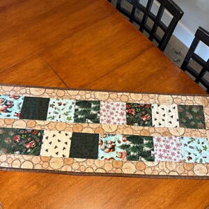 May include: A rectangular table runner with a patchwork design. The runner features alternating squares with various patterns, including forest scenes, snowflakes, and solid colors. The border has a wood-grain pattern.