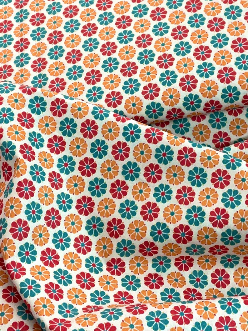 May include: Close-up of a white fabric with a repeating floral pattern. The flowers are in red, teal, and orange, arranged in a grid-like design. The fabric is slightly wrinkled, suggesting it's a textile.