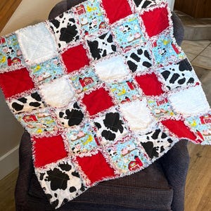 May include: A handmade patchwork quilt featuring a farm animal theme. The quilt has a red, white, black, and blue color scheme. The quilt is made of squares with cow print, red squares, white squares, and a blue farm animal print.