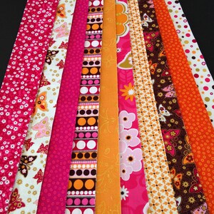 May include: A collection of 10 fabric strips in various patterns and colors, including pink, orange, white, and brown. The strips feature geometric designs, floral patterns, and butterfly motifs.