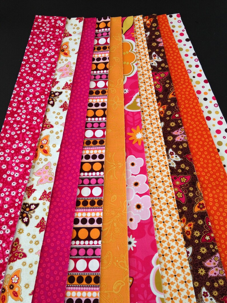 May include: A collection of 10 fabric strips in various patterns and colors, including pink, orange, white, and brown. The strips feature geometric designs, floral patterns, and butterfly motifs.