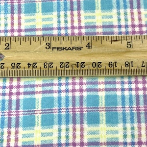 May include: Close-up of a fabric with a plaid pattern in shades of blue, purple, yellow, and white. A wooden ruler with black markings and the text "FISKARS" is placed on top of the fabric. The ruler measures up to 5 inches.