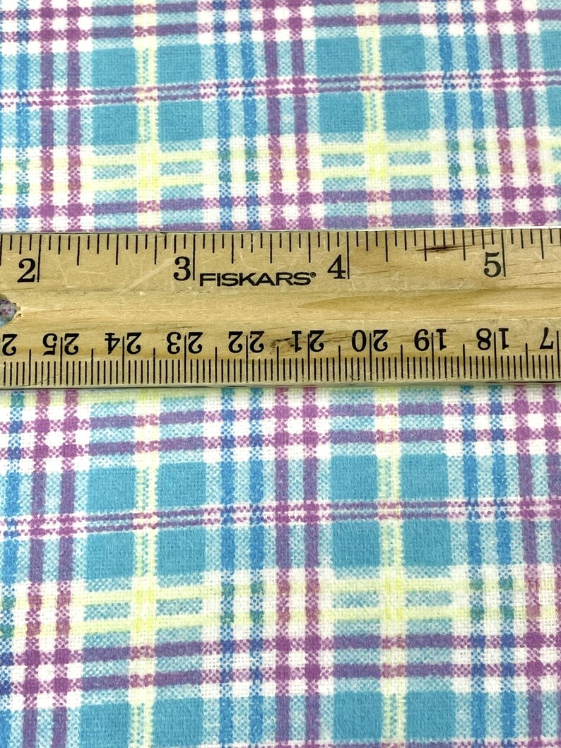 May include: Close-up of a fabric with a plaid pattern in shades of blue, purple, yellow, and white. A wooden ruler with black markings and the text "FISKARS" is placed on top of the fabric. The ruler measures up to 5 inches.