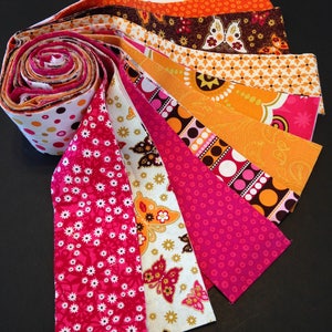 May include: A collection of fabric strips in various patterns and colors, including pink, orange, brown, and white. The strips are arranged in a fan shape, with the ends of the strips overlapping.