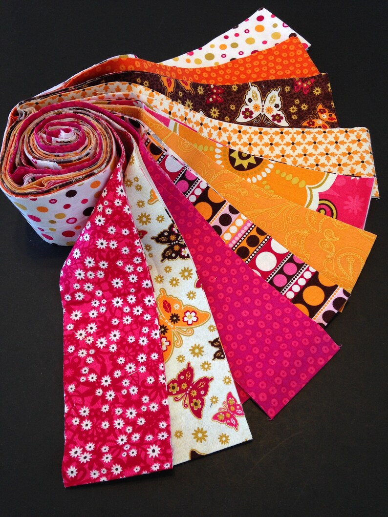 May include: A collection of fabric strips in various patterns and colors, including pink, orange, brown, and white. The strips are arranged in a fan shape, with the ends of the strips overlapping.