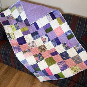 Könnte beinhalten: Ein Patchwork-Quilt mit quadratischen Stoffblöcken in Lila, Rosa, Grün und Weiß. Die Blöcke zeigen florale und gemusterte Designs. Der Rand ist weiß mit einem dünnen Streifenmuster.