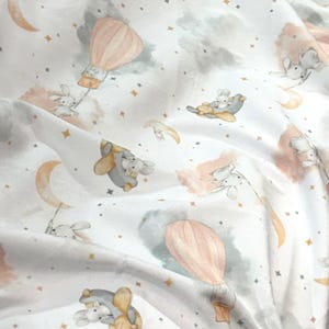 May include: White fabric with a whimsical pattern featuring bunnies, hot air balloons, moons, stars, and clouds in soft pastel colors. The design includes peach, gray, and gold accents, creating a dreamy, celestial theme.