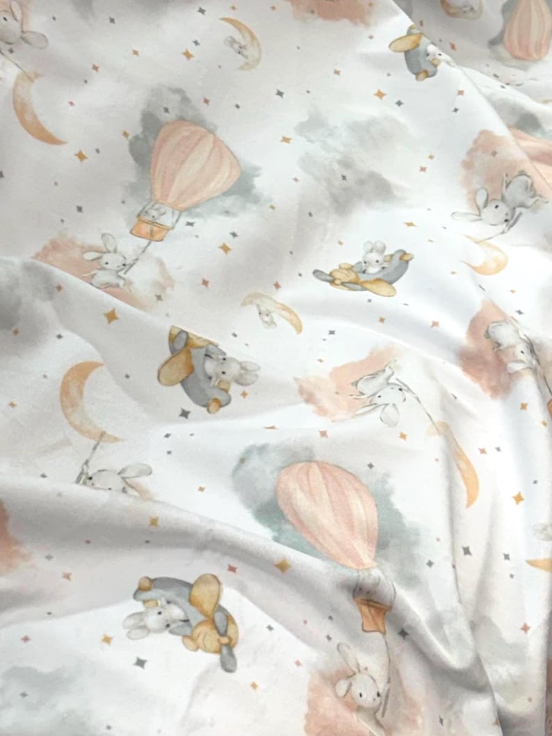 May include: White fabric with a whimsical pattern featuring bunnies, hot air balloons, moons, stars, and clouds in soft pastel colors. The design includes peach, gray, and gold accents, creating a dreamy, celestial theme.