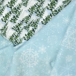 May include: Two pieces of fabric, one with a white background and a repeating pattern of green pine trees covered in snow, and the other a light blue fabric with a white snowflake pattern.
