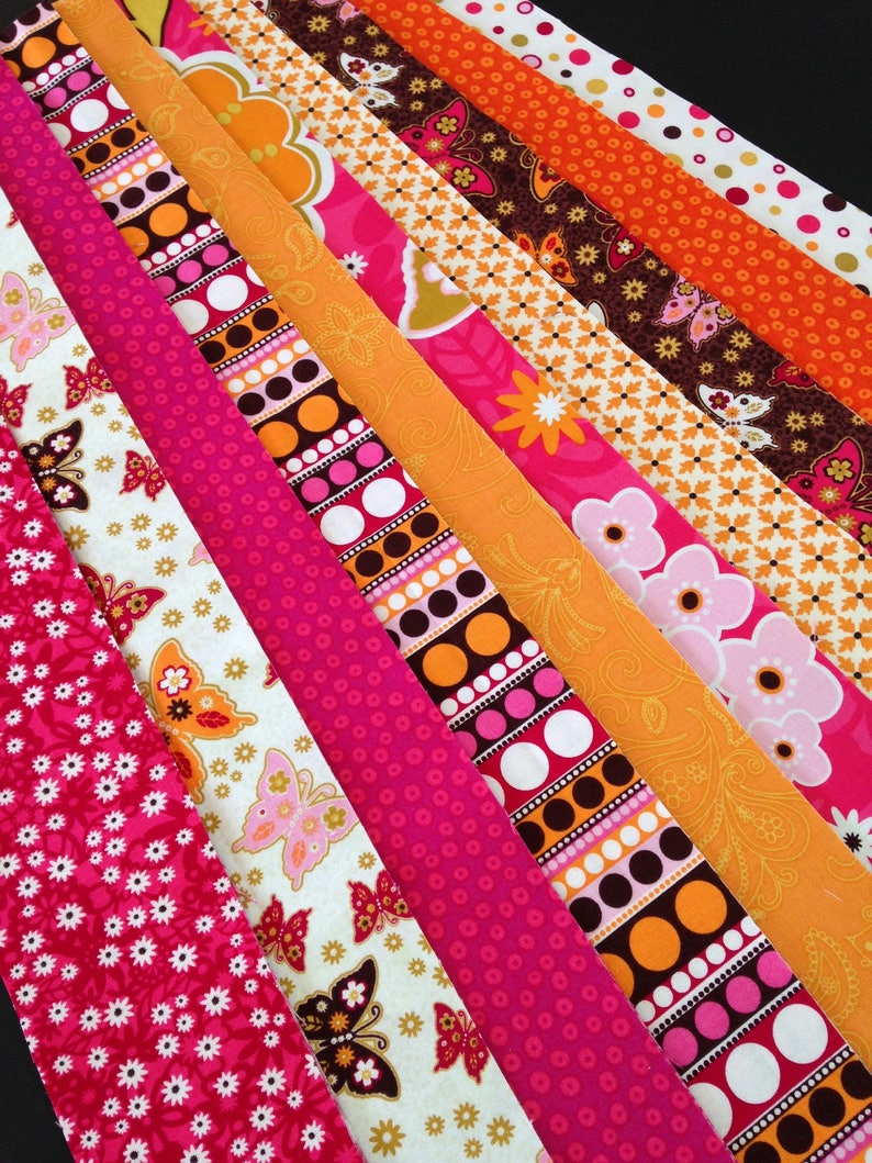 May include: A collection of fabric strips in various patterns and colors, including pink, orange, brown, and white. The strips feature floral, geometric, and butterfly designs.