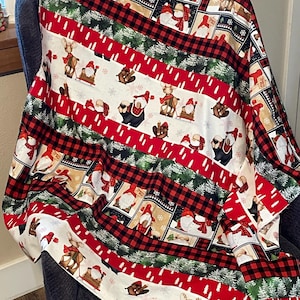 May include: A red, green, and white striped throw blanket with a Christmas gnome pattern. The blanket features gnomes wearing red hats and white beards, along with reindeer, bears, and trees. The blanket is made of soft, cozy fabric and is perfect for adding a festive touch to your home.