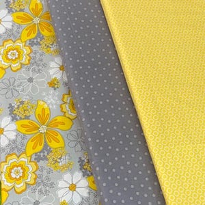 Puede incluir: Primer plano de tres rollos de tela. Las telas presentan estampados florales, de lunares y geométricos en amarillo, gris y blanco. La tela floral tiene grandes flores amarillas con detalles grises y blancos.