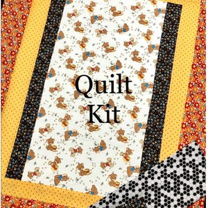 May include: Quilt kit featuring a white fabric with a repeating pattern of bears holding honey pots. The fabric is surrounded by a yellow, black and white, and orange and red border. The text "Quilt Kit" is printed in black on the white fabric.