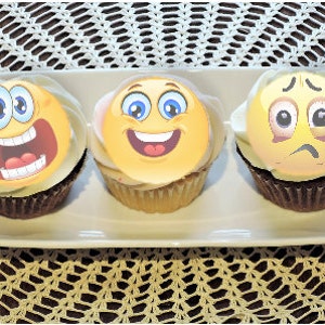 May include: Three cupcakes with emoji faces on a white rectangular plate. The emojis depict expressions of laughter, happiness, and sadness. The cupcakes have white frosting and sit on a brown surface with a white lace doily.