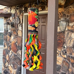 May include: A hanging fall decoration featuring a cylindrical fabric design with a pumpkin and corn graphic. The bottom has colorful, curly ribbons in orange, green, yellow, and black. The decoration is suspended from a black strap.