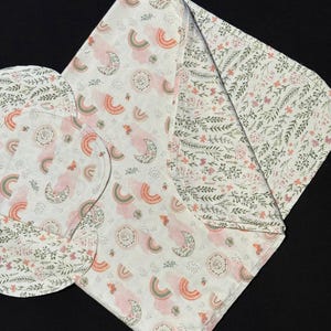 May include: A baby blanket and burp cloths with a whimsical design. The main pattern features rainbows, moons, and floral accents in soft pink, green, and peach hues on a white background. The reverse side of the blanket has a floral pattern.