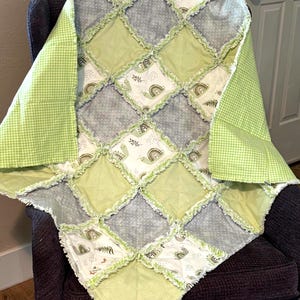 May include: A handmade baby quilt with a patchwork design. The quilt features squares of light green, grey, and white fabric. Some squares have a snail and rainbow pattern. The quilt is trimmed with a frayed edge and a green gingham backing.