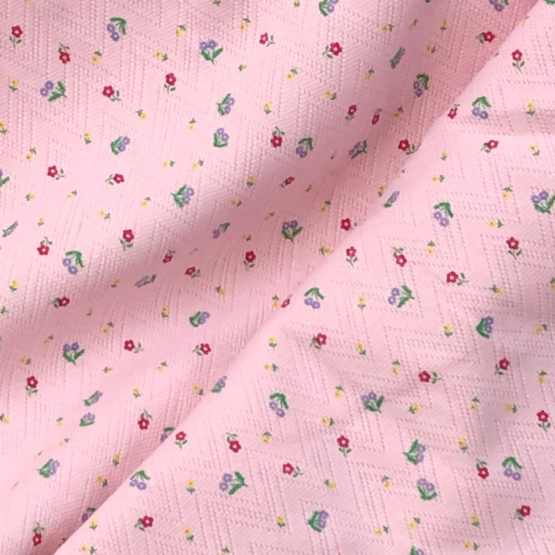 K&ouml;nnte beinhalten: Rosafarbener Stoff mit einem kleinen wei&szlig;en und mehrfarbigen Blumenmuster. Der Stoff hat eine strukturierte, leicht erhabene Oberfl&auml;che.
