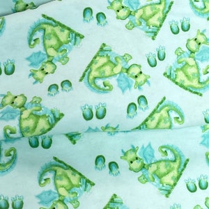 May include: A light blue fabric with a repeating pattern of green dragons and blue footprints. The dragons are facing different directions and have light green wings.