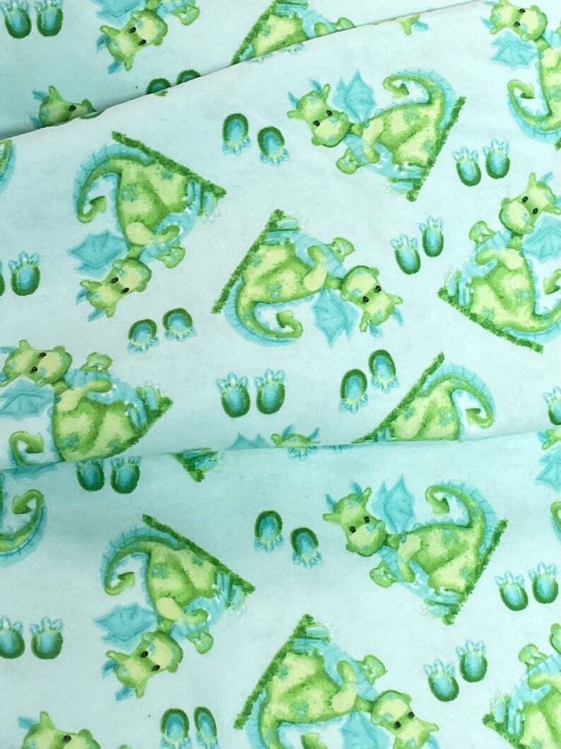 May include: A light blue fabric with a repeating pattern of green dragons and blue footprints. The dragons are facing different directions and have light green wings.
