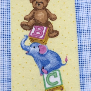 May include: A yellow fabric with a blue and white checkered border. A stack of colorful alphabet blocks with a teddy bear, elephant, and giraffe on top of them.