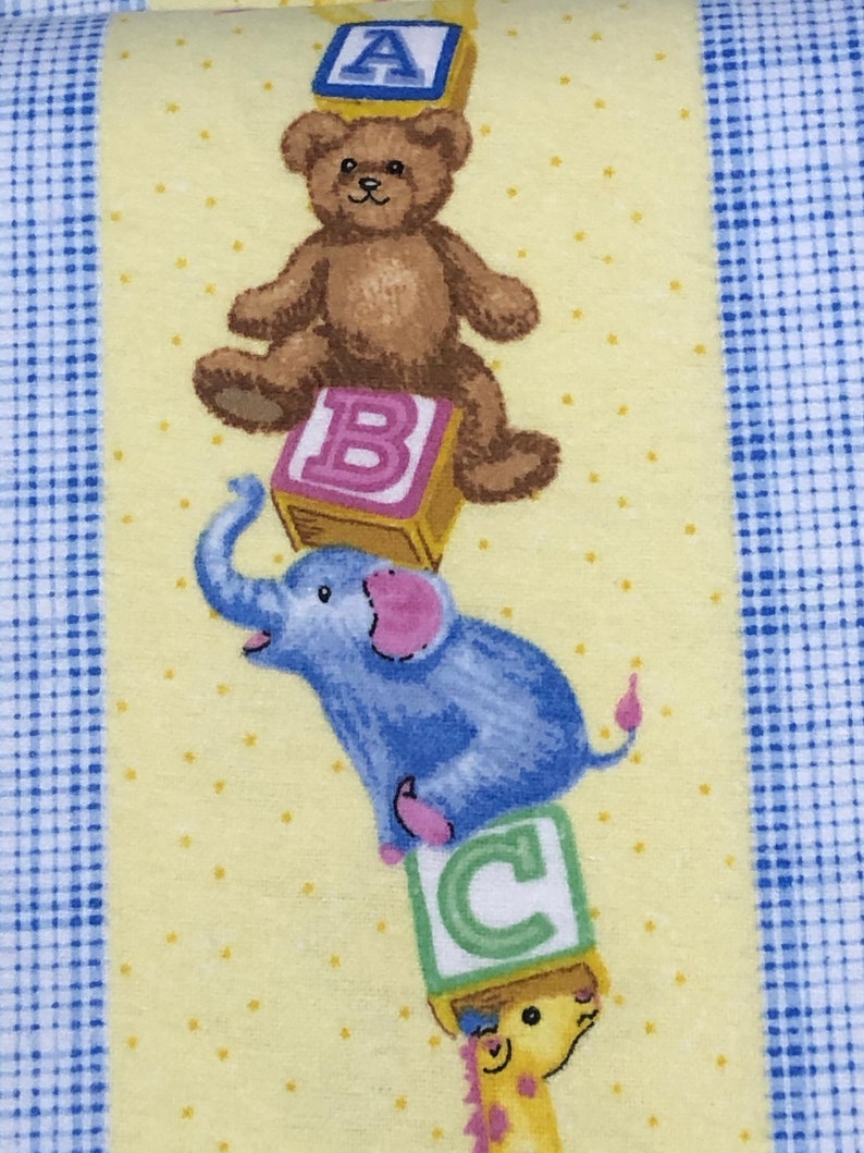 May include: A yellow fabric with a blue and white checkered border. A stack of colorful alphabet blocks with a teddy bear, elephant, and giraffe on top of them.