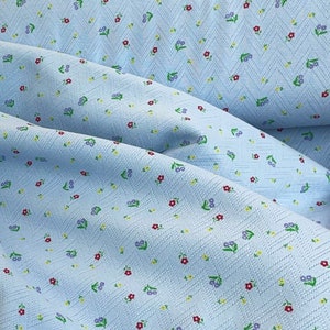 K&ouml;nnte beinhalten: Ein hellblauer Stoff mit einem kleinen Blumenmuster. Der Stoff hat eine strukturierte Oberfl&auml;che und ist diagonal gewebt.