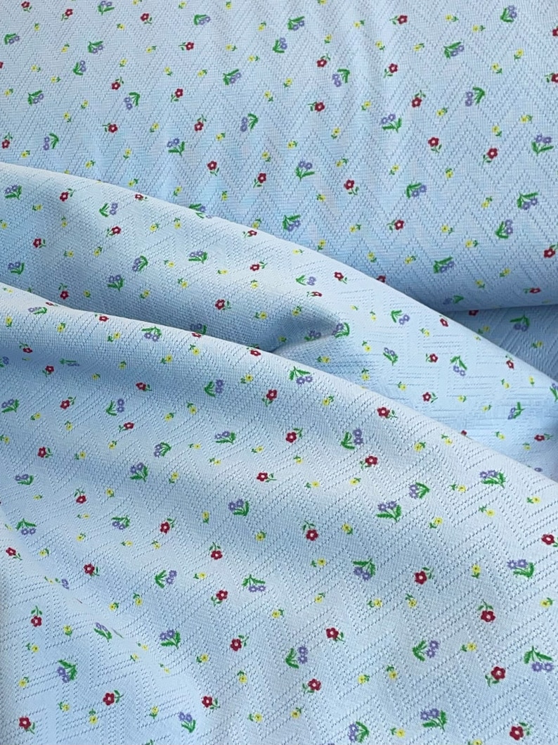 K&ouml;nnte beinhalten: Ein hellblauer Stoff mit einem kleinen Blumenmuster. Der Stoff hat eine strukturierte Oberfl&auml;che und ist diagonal gewebt.
