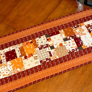 May include: A patchwork table runner with a fall theme. The runner features a mix of orange, burgundy, and cream-colored fabrics with various patterns, including pumpkins, flowers, and geometric shapes. The edges are trimmed with a wavy design and a solid orange border.