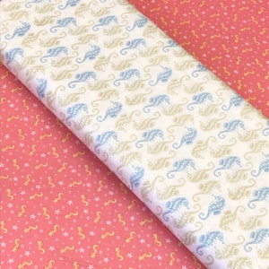 Op de afbeelding: Twee stofstalen met een zeepaardjespatroon. Een staal is wit met blauwe en beige zeepaardjes, de andere is roze met oranje zeepaardjes.