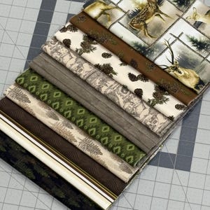 May include: A stack of ten folded fabric swatches featuring various nature-themed patterns. Designs include deer in a forest, brown fabric with pinecones, a cabin scene, solid brown, green geometric shapes, pine trees, and dark forest prints.