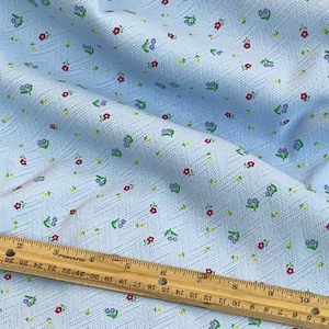 May include: A light blue fabric with a small white, red, and purple floral pattern. The fabric has a textured, woven look.