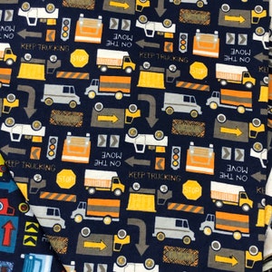 May include: A navy blue fabric with a repeating pattern of construction vehicles, traffic signs, and road markings. The pattern includes yellow construction vehicles, orange traffic cones, and white traffic signs with black text. The text on the signs includes "Keep Trucking", "Move on the", "Stop", and "Detour".