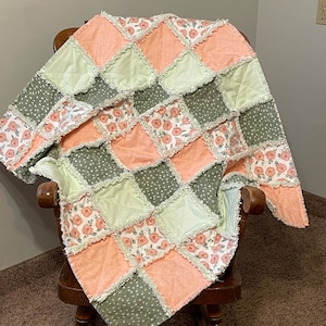 Pode incluir: Um cobertor de bebê em patchwork com um padrão de losangos em tons de pêssego, verde e branco. O cobertor apresenta uma estampa floral com uma borda franjada.