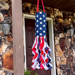 May include: A decorative windsock in patriotic colours. The top section is blue with white stars, and the bottom has red, white, and blue ribbons. It is suspended by a red strap.