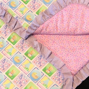 May include: A pink and white baby blanket with a ruffled edge. The blanket has a checkered pattern on one side and a pink background with white polka dots on the other side. The blanket features a repeating pattern of baby animals and the word "BABY" in a blue and yellow font.