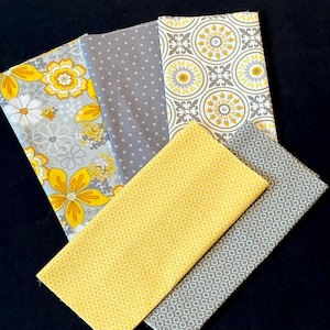 May include: A collection of folded fabric pieces in various patterns and colors. Designs include yellow flowers on gray, gray polka dots, yellow and gray geometric shapes, and solid yellow and gray fabrics. The fabrics are arranged on a dark surface.
