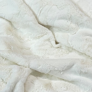 May include: Close-up of a white, plush blanket with a textured, fur-like appearance. The fabric is folded, creating shadows and highlights that emphasize the soft, luxurious texture. The blanket appears to be made of a thick, warm material.