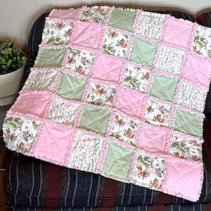 May include: A handmade patchwork quilt with a charming woodland theme. The quilt features squares of pink, green, and floral fabric with a ruffled edge. The floral fabric has a deer and botanical design.