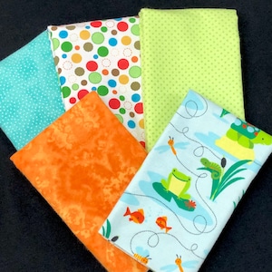 Könnte beinhalten: Fünf Stoffquadrate in verschiedenen Mustern. Ein Quadrat ist orange mit einem Marmormuster. Ein Quadrat ist hellblau mit weißen Punkten. Ein Quadrat ist weiß mit roten, braunen und blauen Punkten. Ein Quadrat ist hellgrün mit weißen Punkten. Ein Quadrat ist hellblau mit einem Muster aus grünen Fröschen, Schildkröten und Libellen.