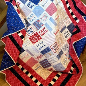 May include: A patchwork quilt featuring a baseball theme with red, white, and blue fabric squares. The quilt displays text including "Play Ball," "Fast Ball," "Home Run," "Strike Out," "Grand Slam," "Batter Up," and "Slugger" in various fonts.