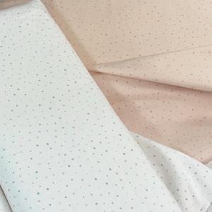 Könnte beinhalten: Zwei Stoffstücke, eines weiß und eines pfirsichfarben, sind zu sehen. Beide Stoffe sind mit kleinen, gleichmäßig verteilten Sternen in Rosa-, Blau- und Grüntönen gemustert. Der weiße Stoff ist gefaltet, während der pfirsichfarbene Stoff ausgebreitet ist.