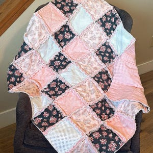 Puede incluir: Un edredón patchwork hecho a mano en tonos rosa, blanco y gris oscuro, sobre una silla. El edredón presenta una variedad de patrones florales y de lunares, con un borde con volantes. Las dimensiones del edredón no son visibles.