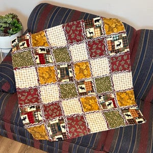Peut inclure: Une courtepointe patchwork sur le thème rustique, avec des carrés de différents motifs et couleurs. La courtepointe incorpore des tissus avec des motifs de feuilles, d'ours et d'orignaux dans des tons de rouge, d'or, de vert et de crème. La courtepointe est posée sur un canapé rayé.