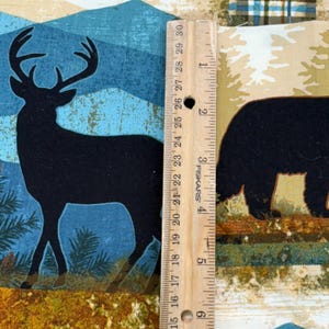 May include: Fabric with a nature-themed design featuring a black silhouette of a deer with antlers, a bear, and mountain scenery. The fabric is measured with a wooden ruler, showing a length of approximately 6 inches.